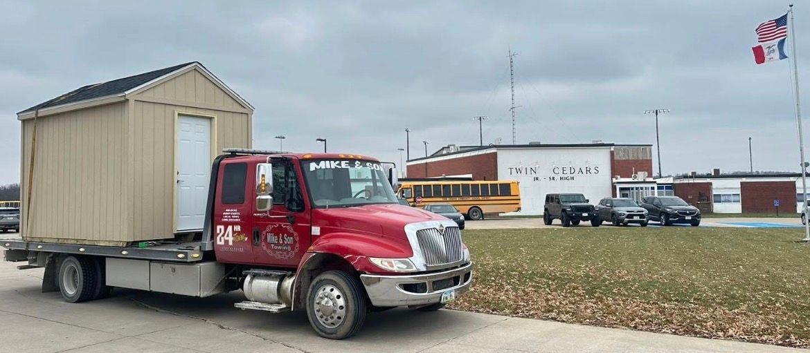 A red truck transporting a shed. In the background, a school building and a flag.