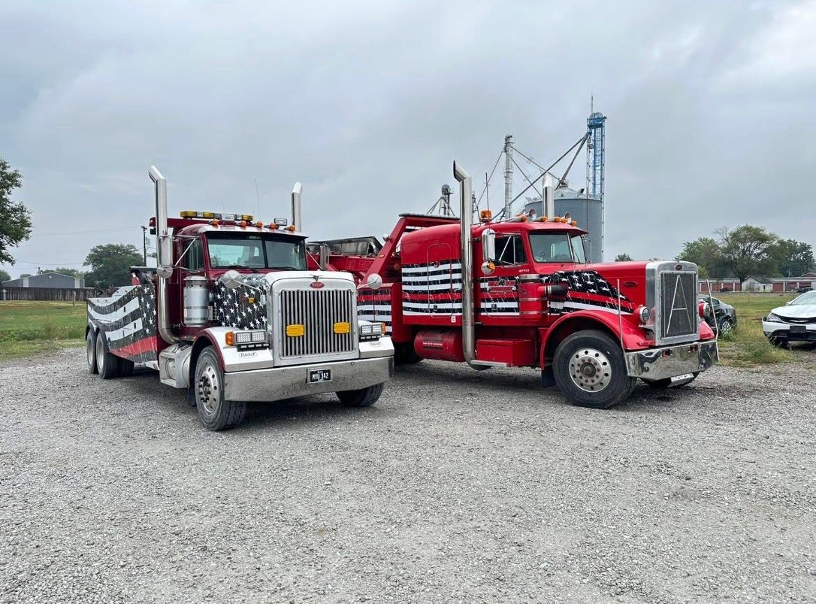 Two red and silver tow trucks parked on gravel; a silo in the background under a cloudy sky.