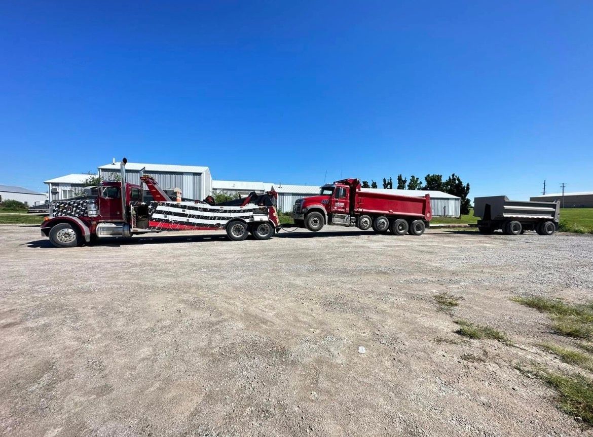 Red tow truck towing a red dump truck and a smaller dump trailer on a gravel lot under a blue sky.