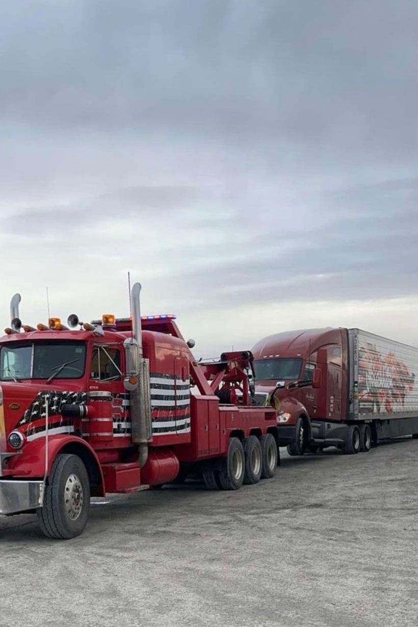 Red tow truck pulling a semi-truck with a red trailer against a cloudy sky.