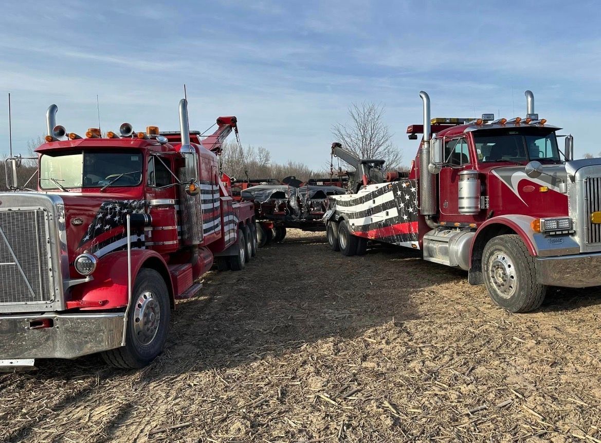 Two red tow trucks towing a damaged vehicle on a gravel lot under a blue sky.
