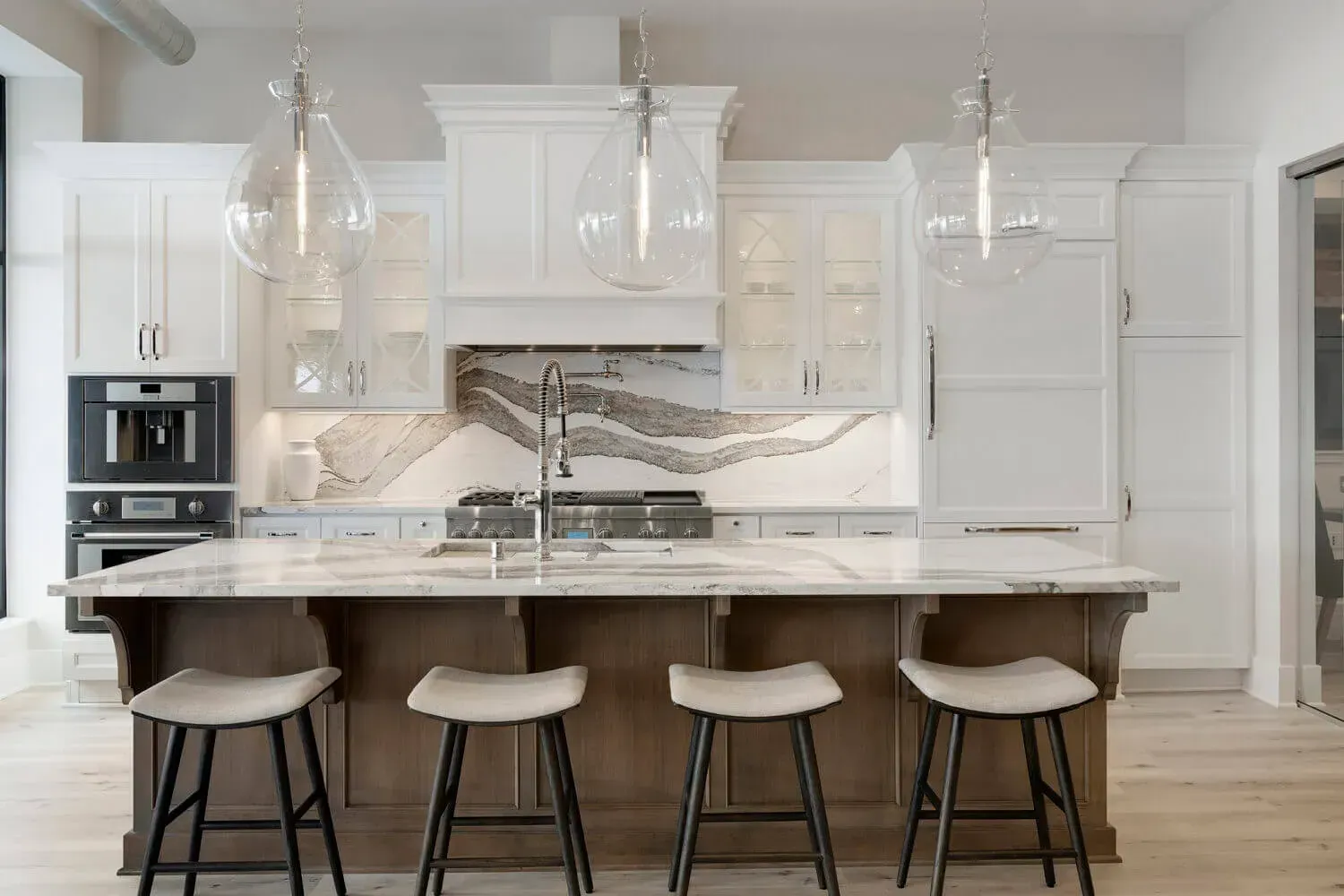 Elegant white kitchen with marble countertops, wood island, glass pendant lights, and bar stools.