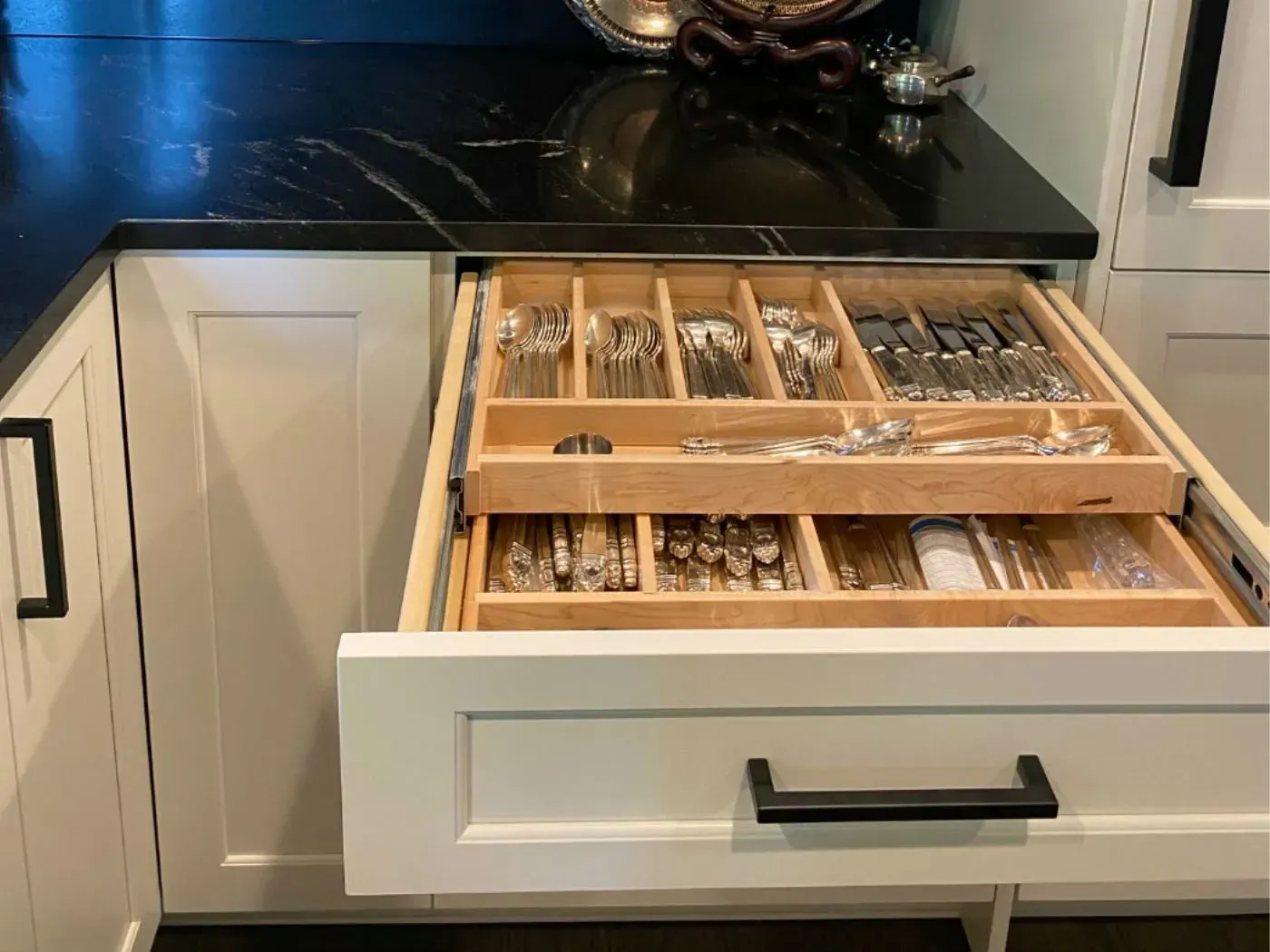 Open white kitchen drawer with two wooden silverware trays. Black countertop and cabinet hardware.
