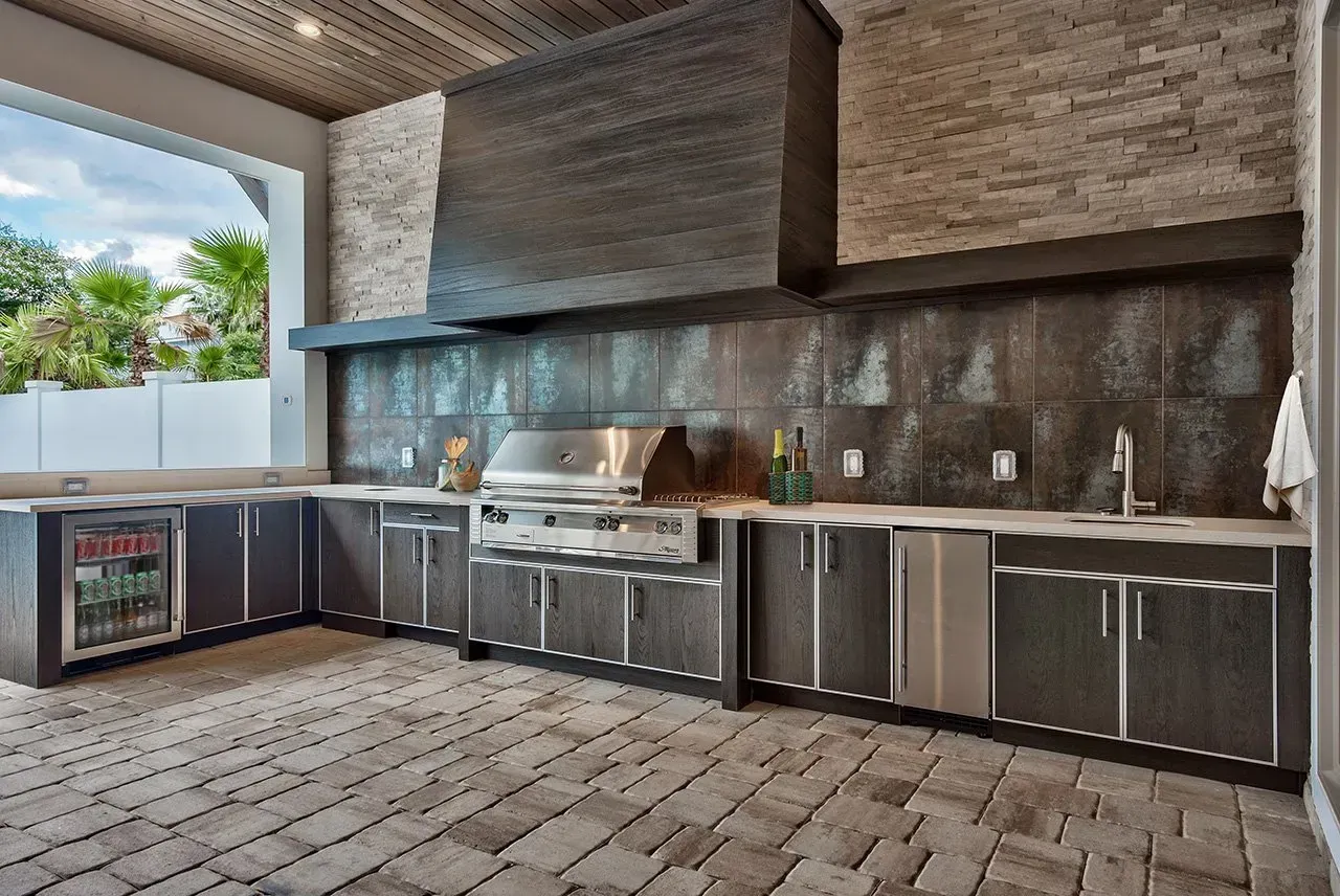 Outdoor kitchen with dark cabinetry, stainless steel grill, brick wall, and stone flooring.