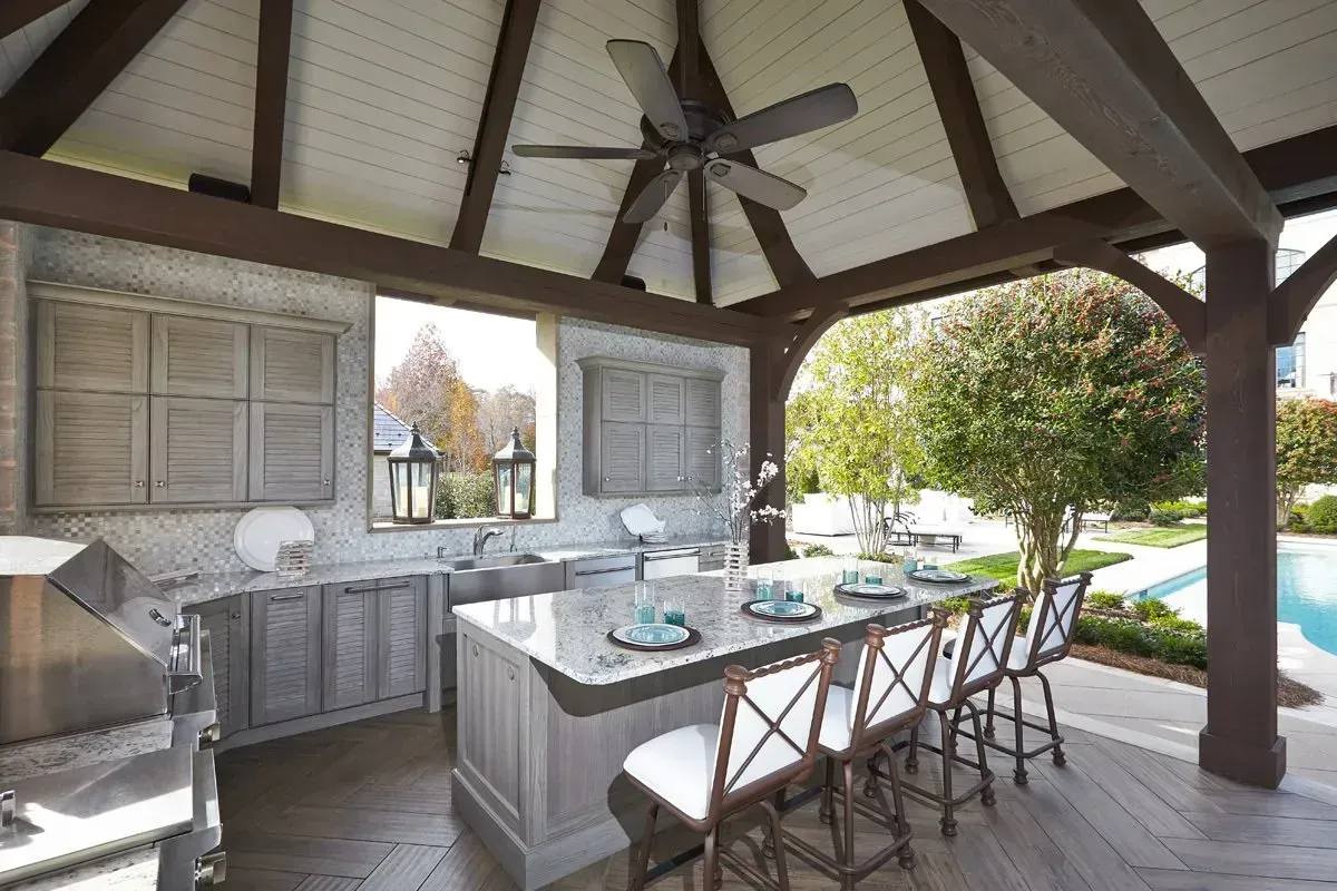 Outdoor kitchen under a wooden gazebo with a grill, cabinets, island, and seating overlooking a pool.