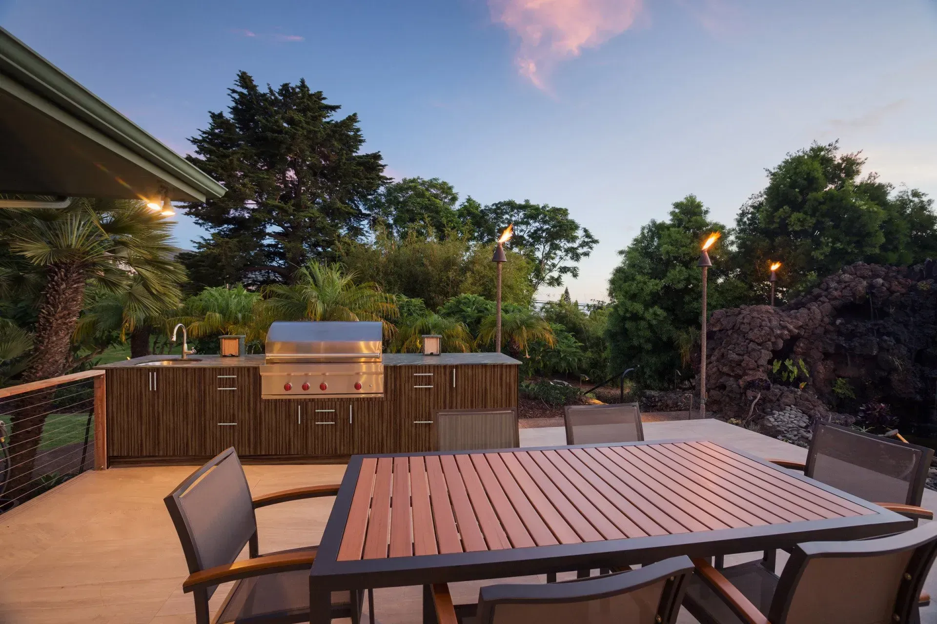 Outdoor patio with grill and dining table, surrounded by lush trees under a twilight sky.