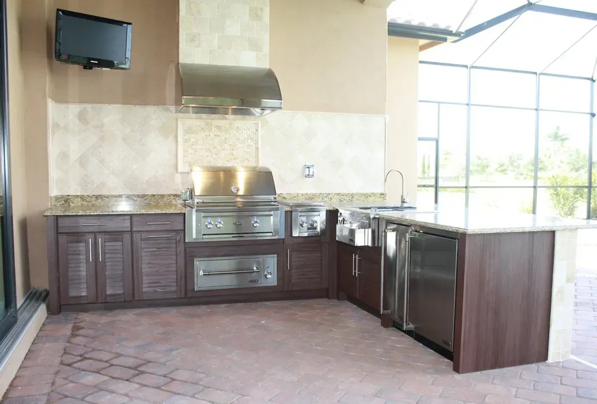 Outdoor kitchen with stainless steel appliances, dark wood cabinets, and beige countertop, under a screened enclosure.