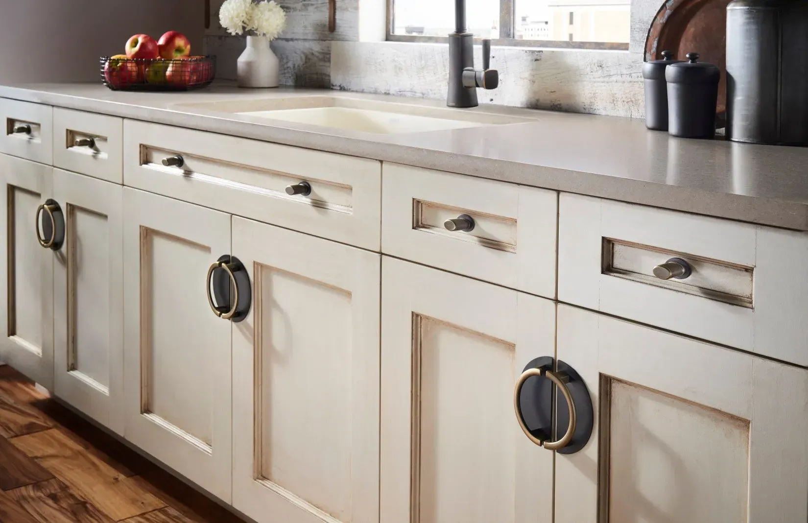 Cream-colored kitchen cabinets with dark hardware against a light countertop, sink, and window.
