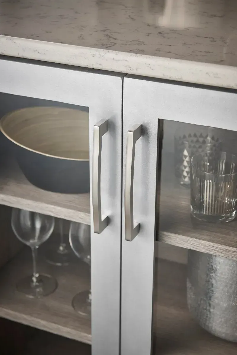 Close-up of a white cabinet with glass doors, showing dishware and glasses inside. Silver handles.