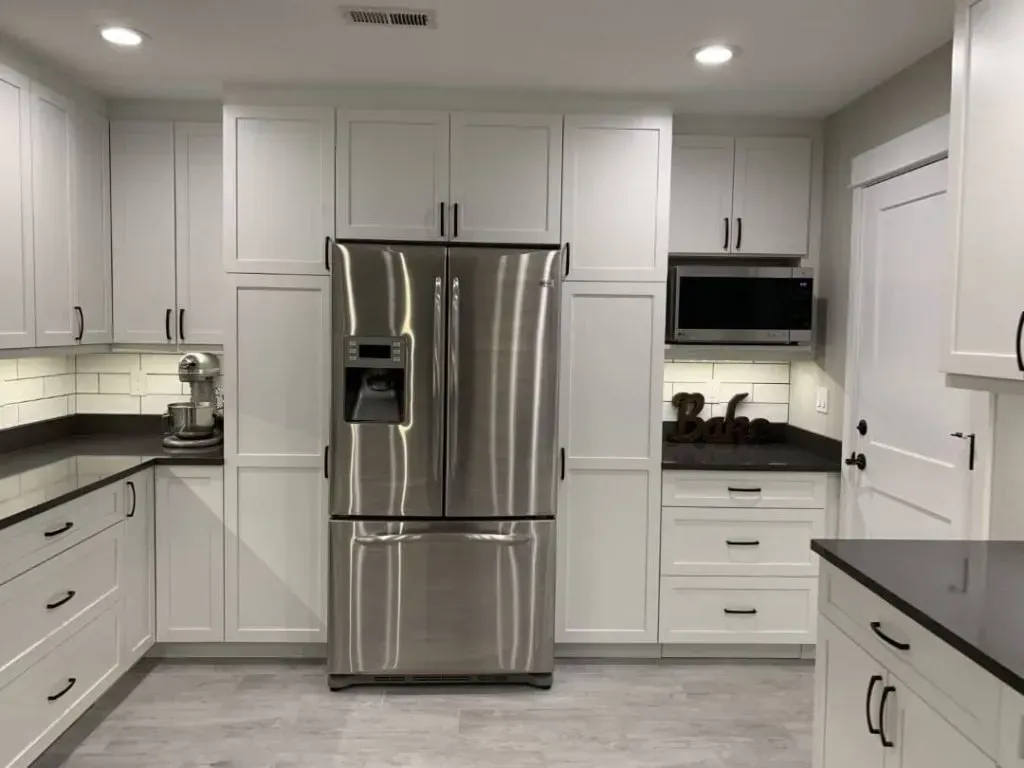 White kitchen with stainless steel refrigerator, white cabinets, dark countertops, and light gray flooring.