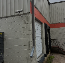 Gray industrial building with red trim, wall-mounted security camera, and exterior stairway.