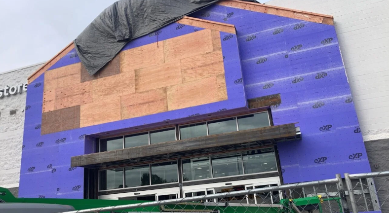Building under construction wrapped in purple insulation, with exposed wood framing and a black tarp on the roof.