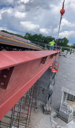Red steel beam being hoisted on a rooftop construction site with workers and concrete blocks below