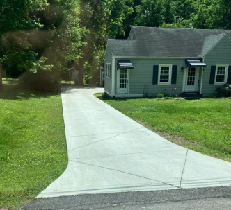 Concrete driveway leading to a small gray house beside a grassy lawn and trees.