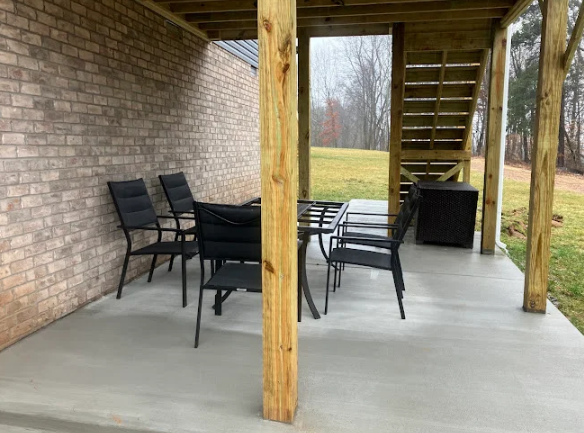 Covered patio with black chairs and table on a concrete floor beside a brick wall and wooden posts