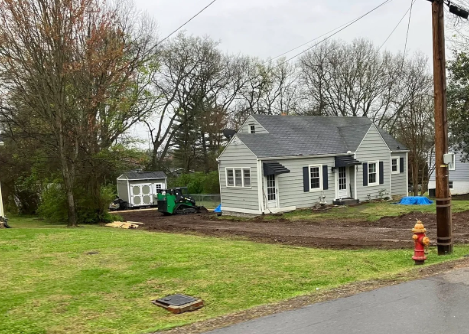 Small gray house with white trim in a yard, with a fire hydrant and roadside in the foreground.