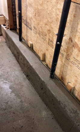Concrete curb along plywood wall with black metal posts in a narrow construction area.