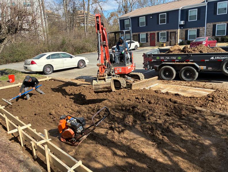Workers and excavator digging a muddy foundation beside a suburban house, with truck and car nearby