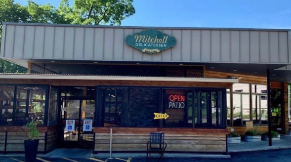 Small storefront with green sign, large windows, and an open “OPEN” neon sign under a covered walkway.