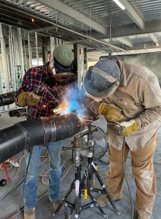 Two welders working on metal pipes in an industrial interior, with bright sparks flying.