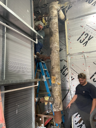 Two workers on ladders inside a partially finished room, handling a large vertical pipe or duct.