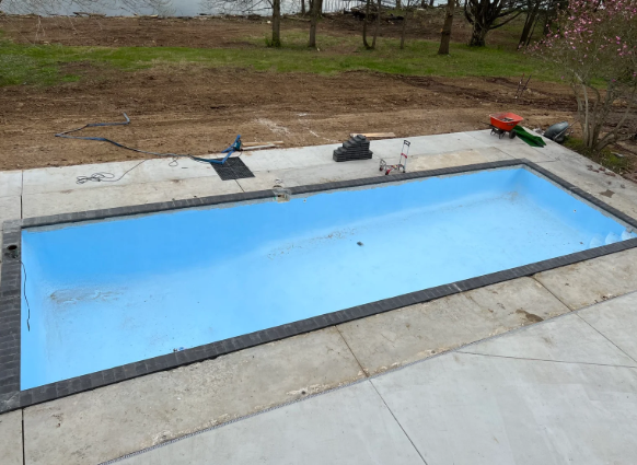 Empty rectangular blue swimming pool beside a concrete patio and dirt yard