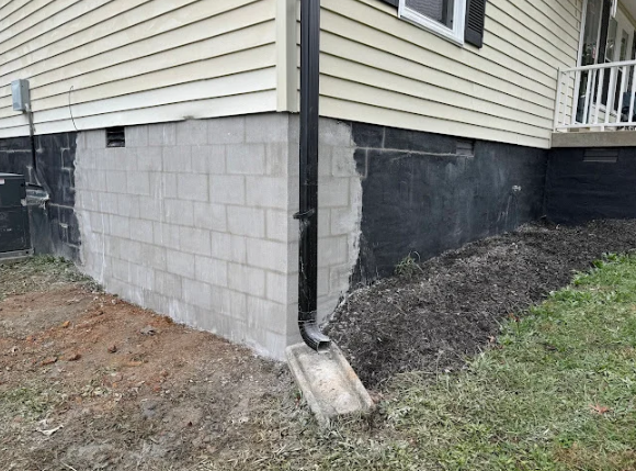 House corner with beige siding, gray cinderblock foundation, and a downspout draining to the ground