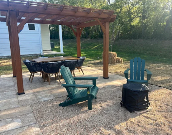 Backyard patio with wooden pergola, dining table, and three colorful chairs on a paved and gravel area.