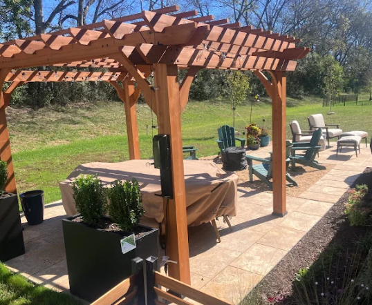 Wooden pergola on a sunny patio with outdoor seating, potted plants, and a covered table near the garden.