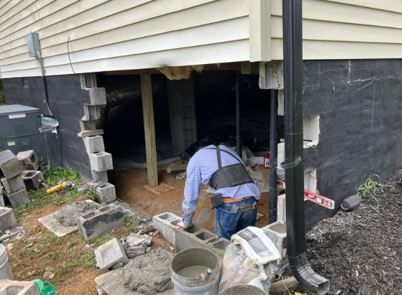 Worker repairing damaged house foundation under siding, with debris and concrete blocks nearby