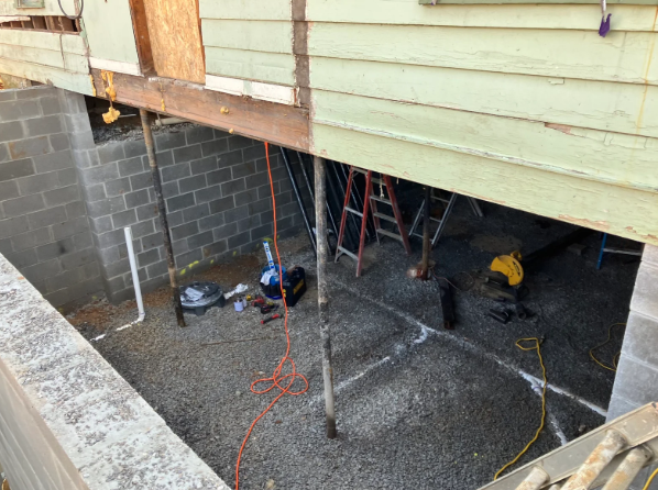 Under-house construction area with cinder block walls, tools, ladders, and yellow power equipment on dirt ground