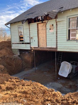 House with severe foundation damage and exposed crawlspace, with dirt pile and ladder beneath the porch