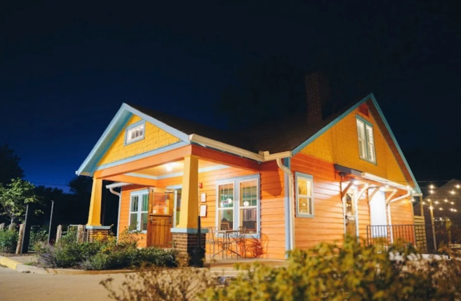 Well-lit orange house with a porch at night, surrounded by a fence and trees.
