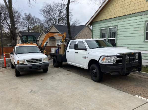 Two white utility trucks parked beside a house, with an excavator and construction cones in the background.