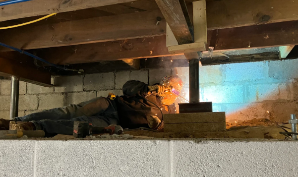 Person lying low under a wooden structure with sparks flying from a tool, suggesting construction or repair work