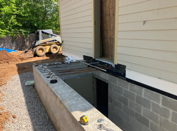 Partially built concrete foundation beside a beige house, with construction equipment in the background