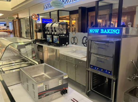 Bakery counter with display cases and a blue “NOW BAKING” sign in a mall food court