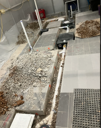 Construction site walkway with gray concrete blocks, rubble, trench, and drainage grates along a wall