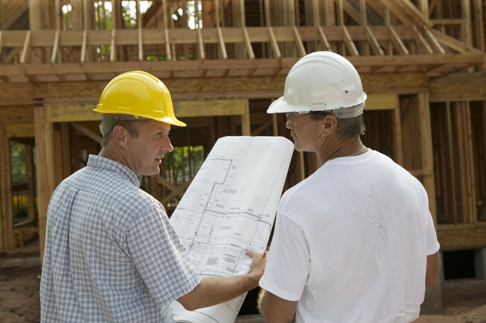 Two construction workers in hard hats reviewing blueprints at a wooden house frame