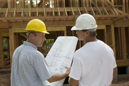 Two construction workers in hard hats reviewing blueprints at a wooden house frame