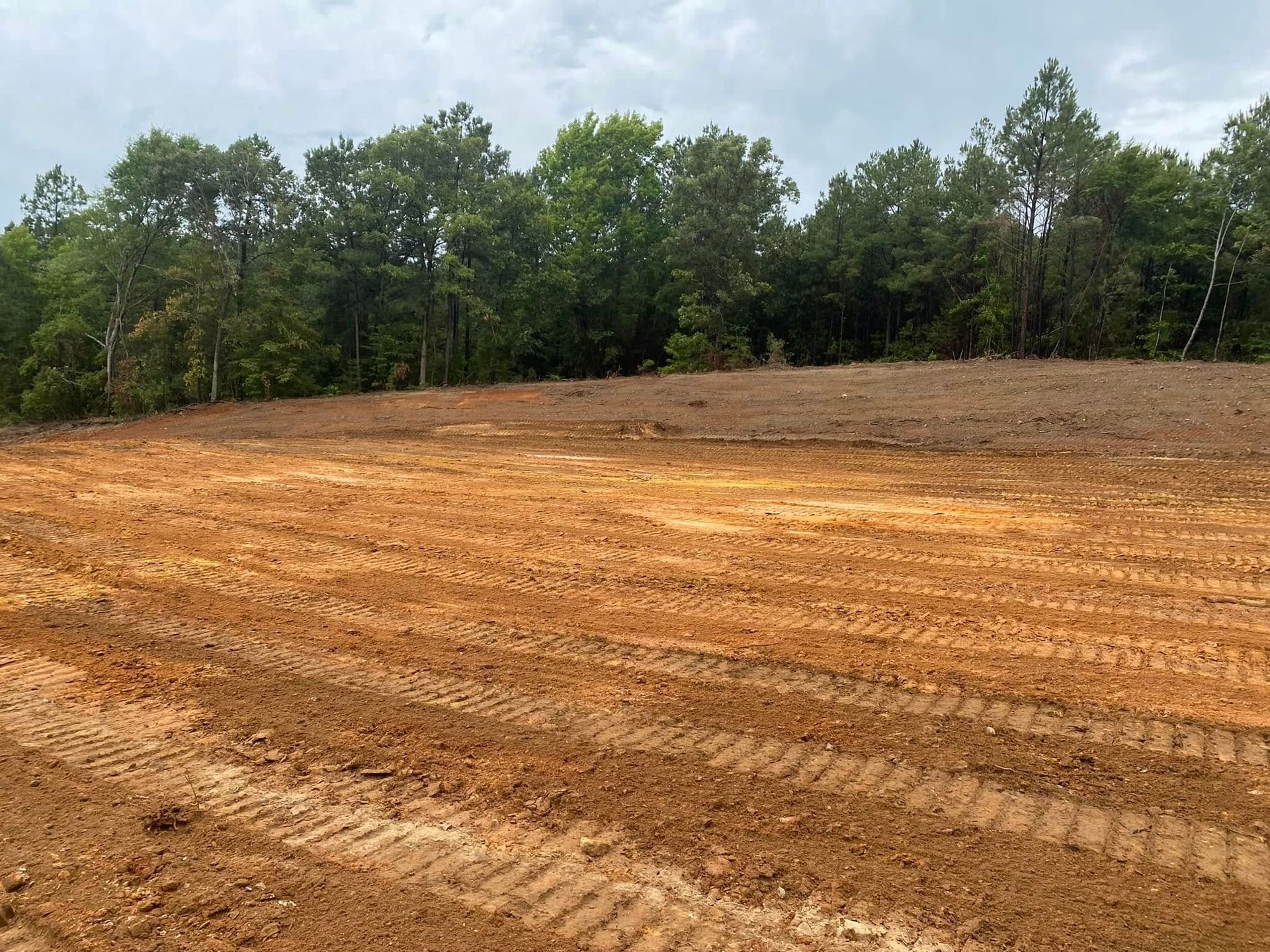 Freshly graded orange-brown dirt lot beside a green tree line under a cloudy sky