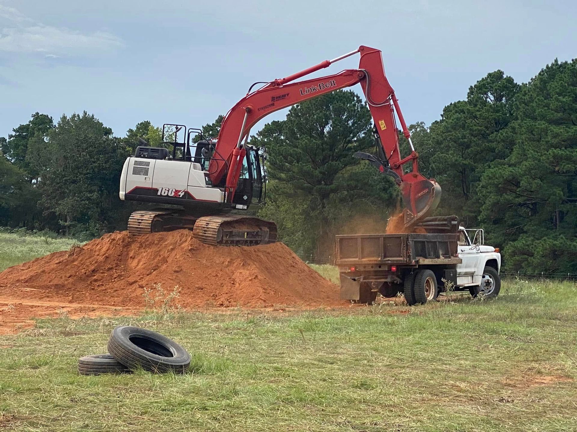 Excavator loading red dirt into a dump truck on a grassy worksite, with tires in the foreground.