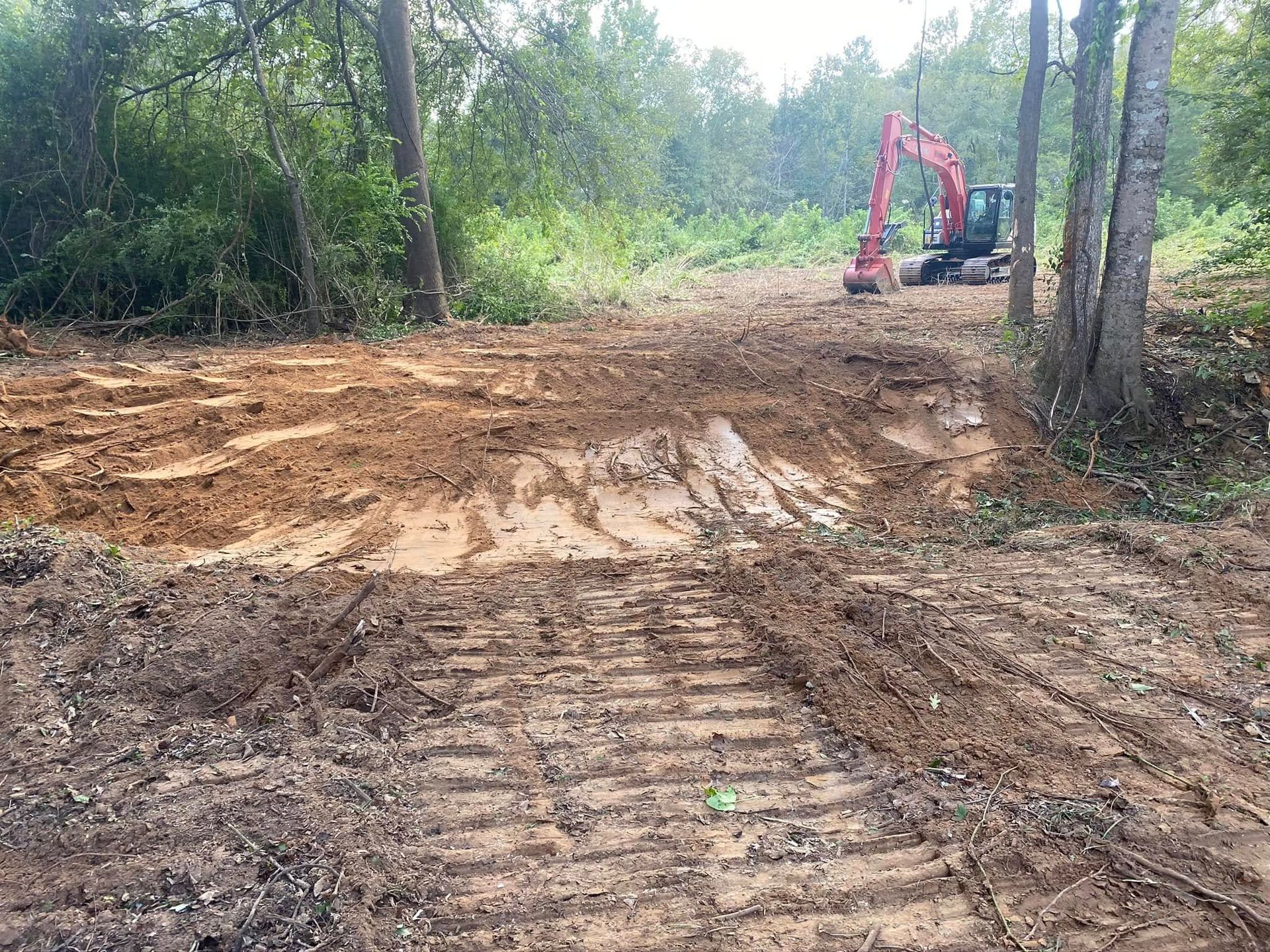 Dirt road under construction in a wooded area, with a small excavator in the distance.