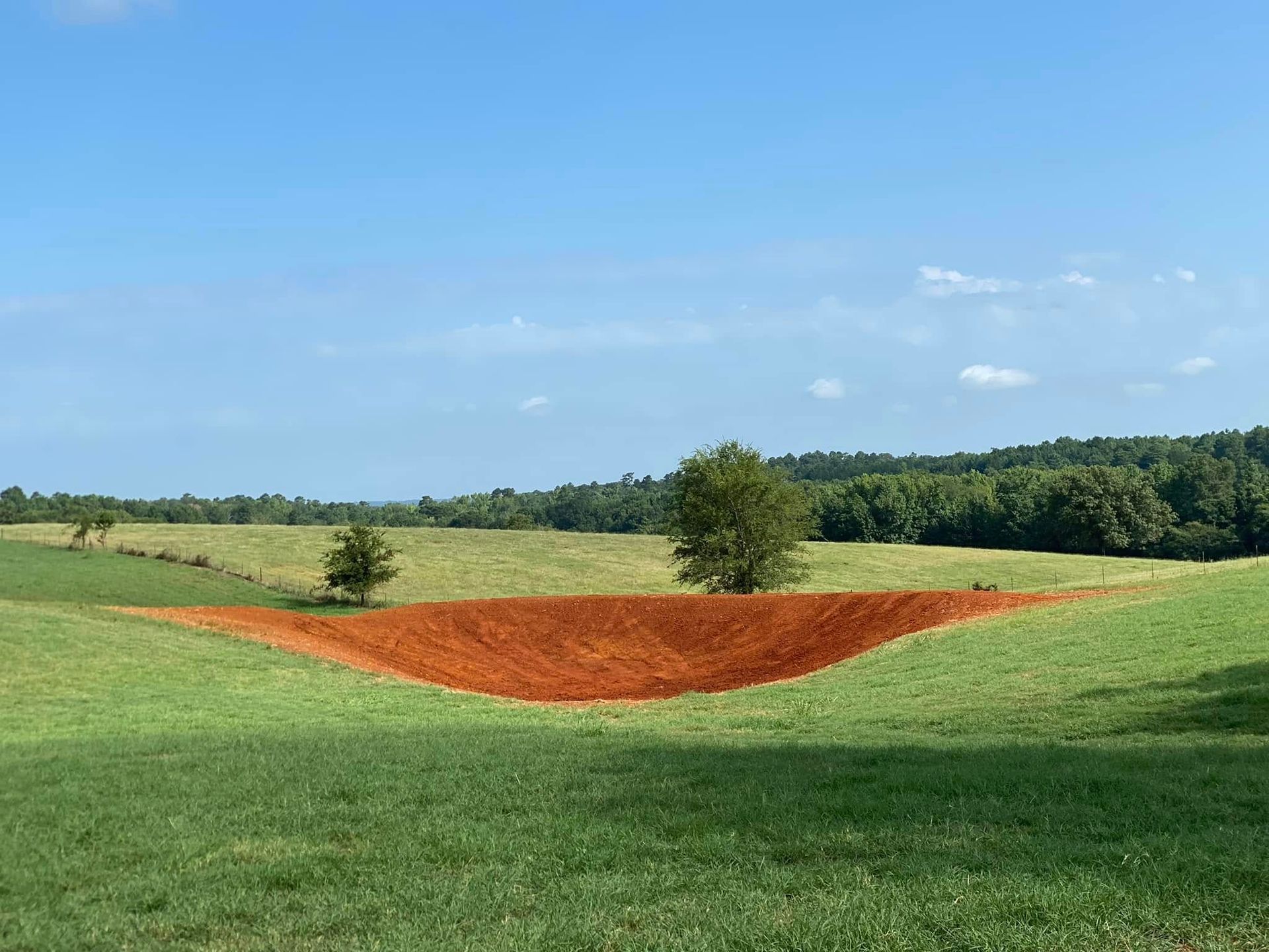 Reddish-brown bare patch in a green field under a blue sky with scattered trees