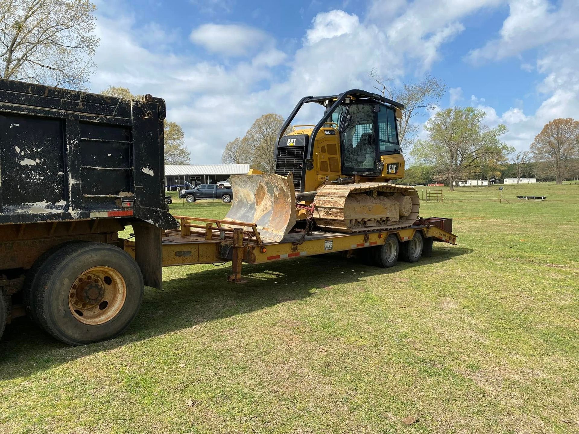 Yellow bulldozer on a flatbed trailer beside a black dump truck in a grassy field under a partly cloudy sky
