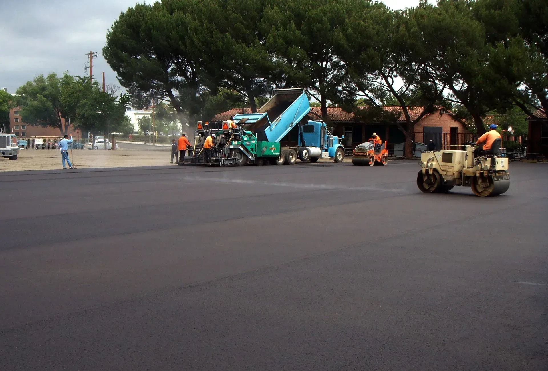 Road workers paving a street with machinery and a small vehicle under trees