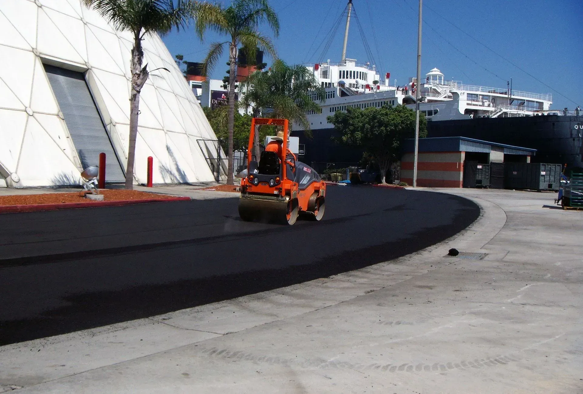 Orange forklift paving a curved black road beside white sails and a harbor