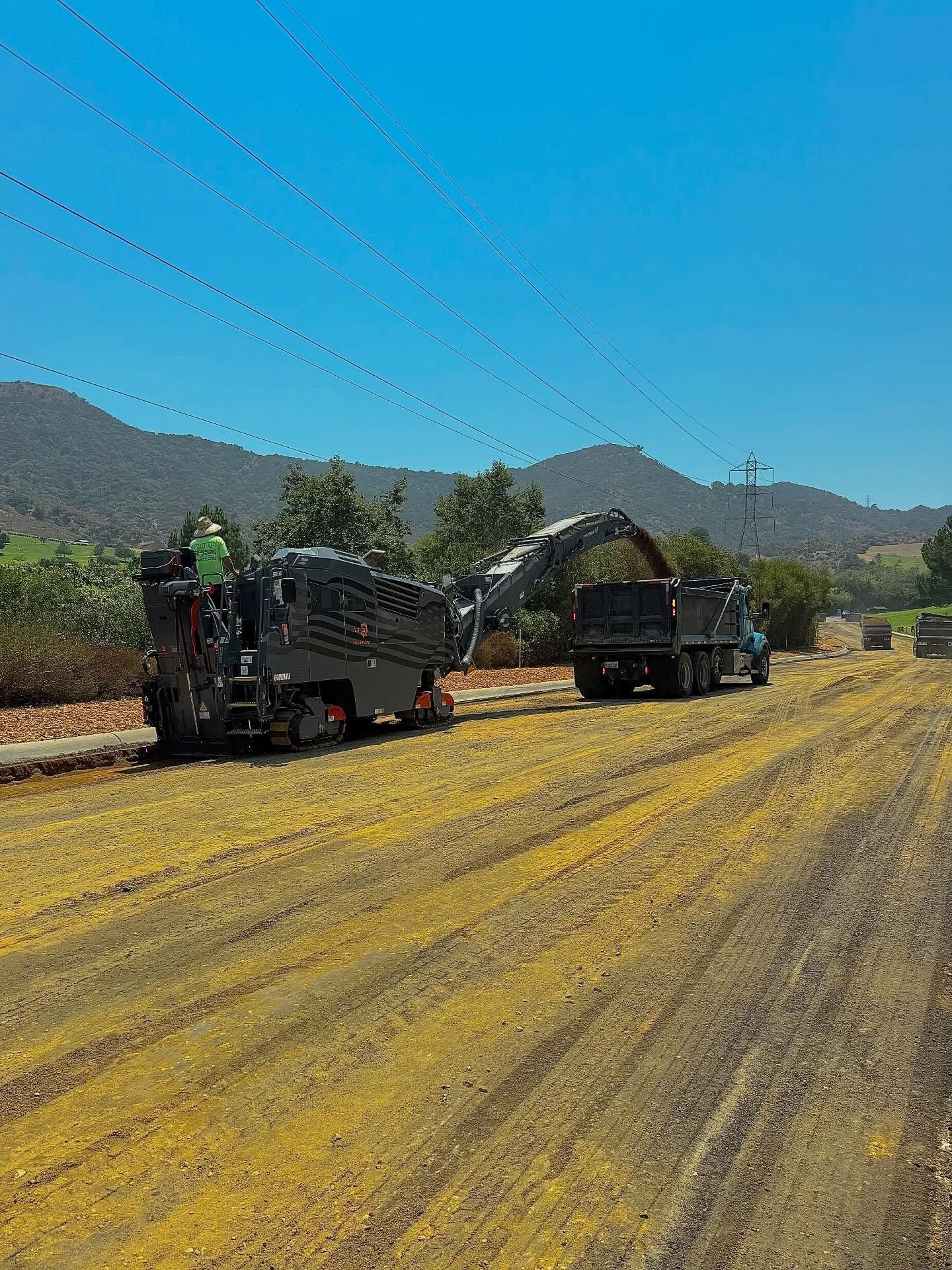 Two road machines on a yellow dirt road under a clear blue sky, with mountains in the background