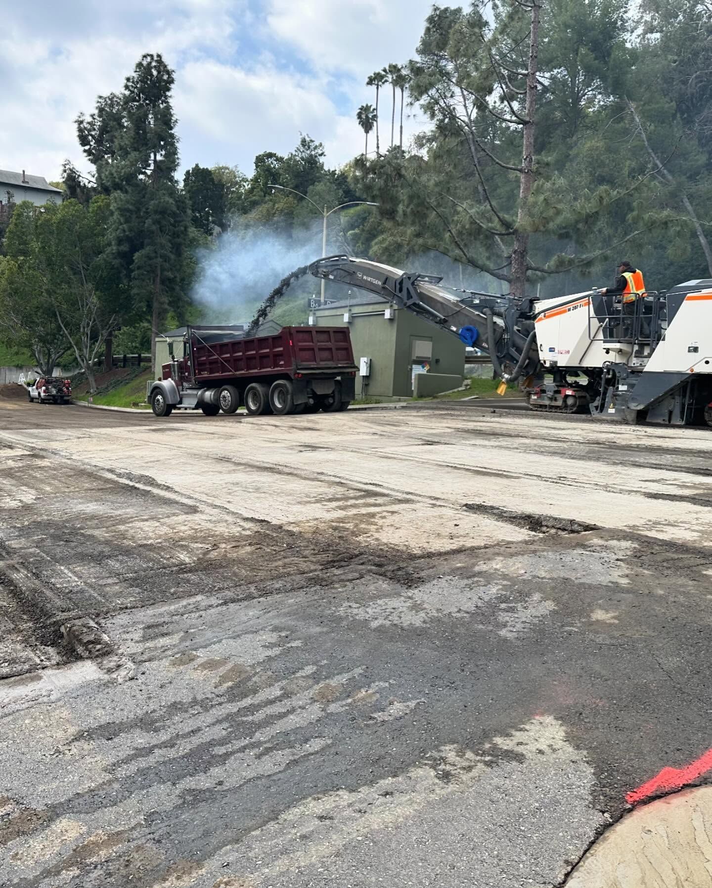 Road paving machine dumping asphalt into a red truck on a roadside worksite, with smoke rising nearby