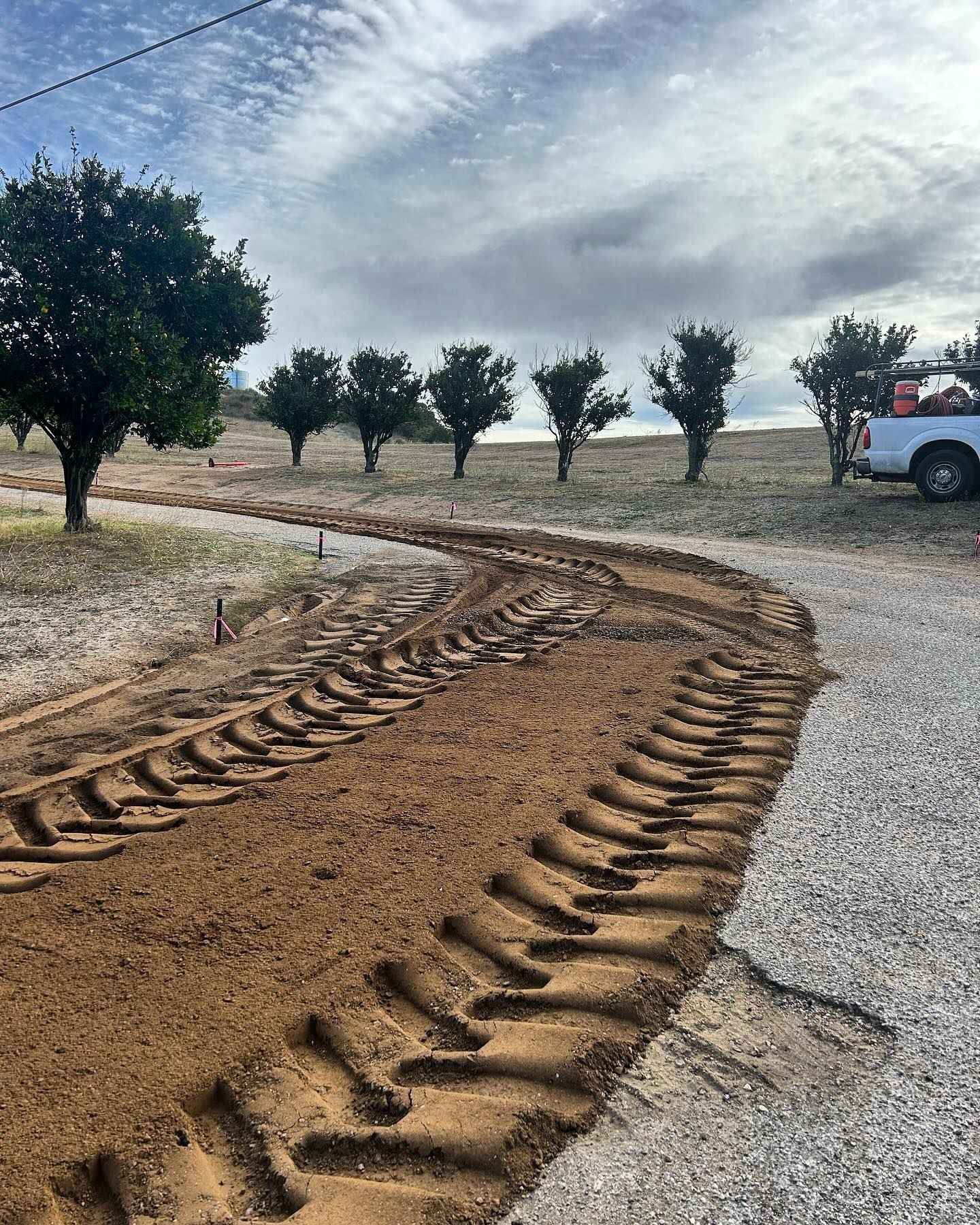 Curving tire tracks in gravel leading past small trees under a cloudy sky, with a white truck at right.